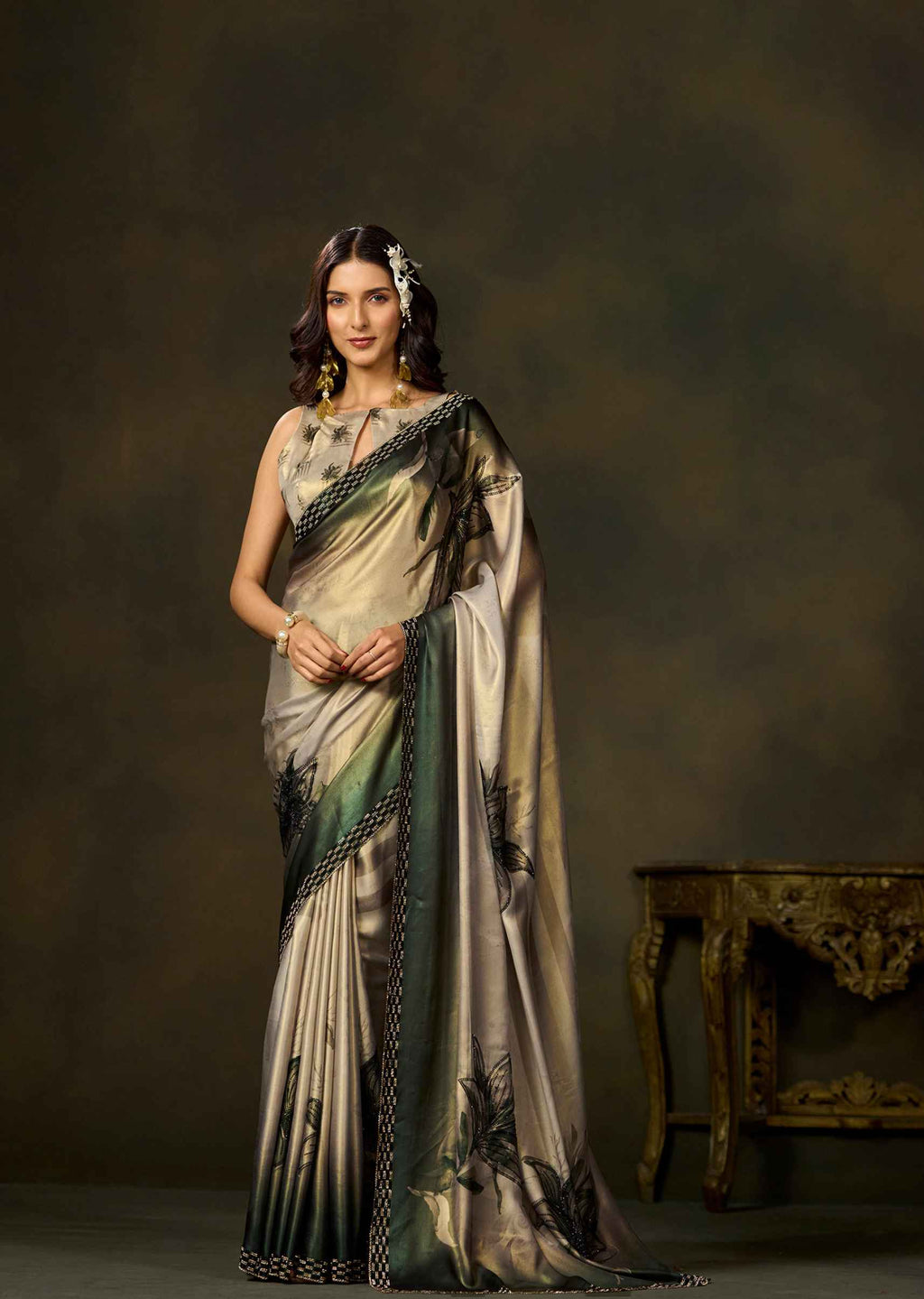 A woman models a traditional Indian saree with a beige and green floral pattern, set against a dark brown studio backdrop with an ornate table on the right.