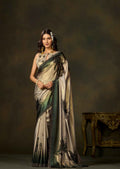 A woman models a traditional Indian saree with a beige and green floral pattern, set against a dark brown studio backdrop with an ornate table on the right.