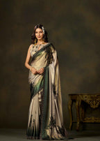 A woman models a traditional Indian saree with a beige and green floral pattern, set against a dark brown studio backdrop with an ornate table on the right.