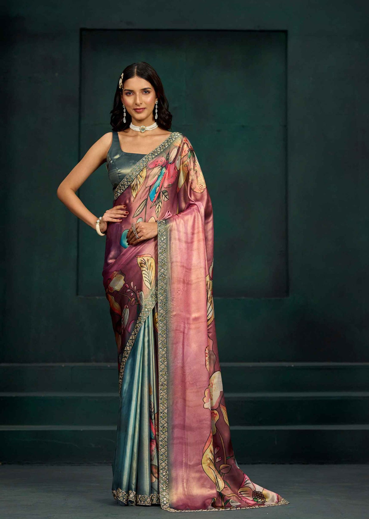 A woman models a traditional Indian saree with a metallic teal-blue body, a pink and gold floral-patterned pallu, and an embellished gold border, paired with a matching metallic teal-blue sleeveless blouse.