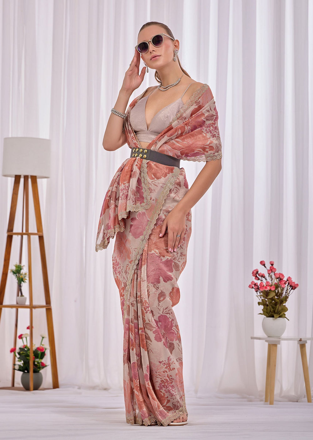 A woman modeling a draped floral saree with a pink and beige floral pattern, styled with a dark belt and sunglasses, standing in front of white curtains with decorative plants and a floor lamp.