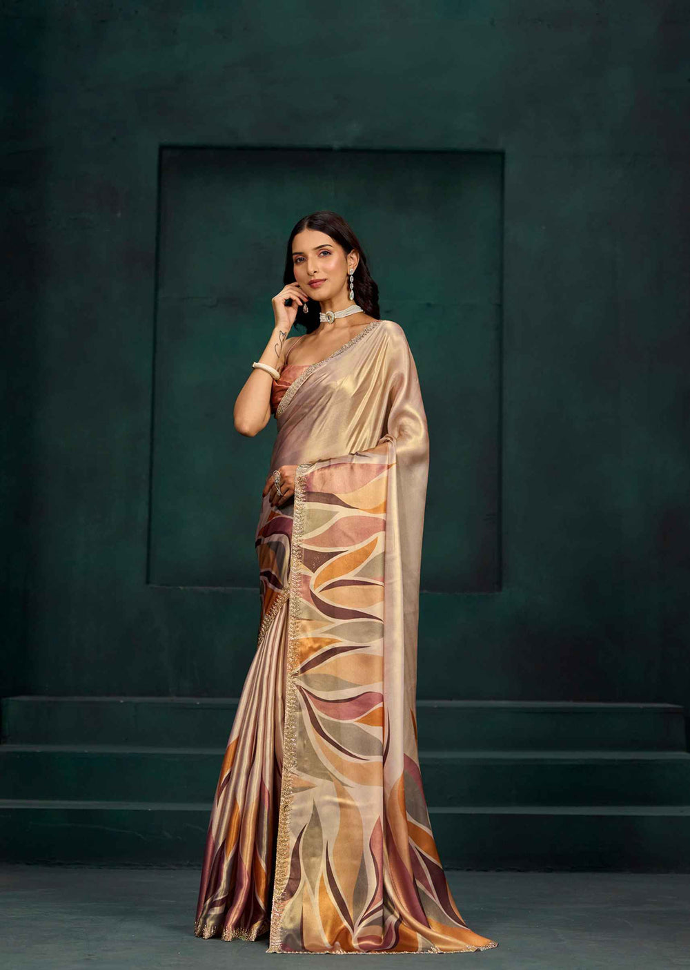 A woman models a draped saree with a metallic champagne-gold base and an abstract leaf-like pattern in bronze, brown, and orange tones, set against a dark green textured studio backdrop.