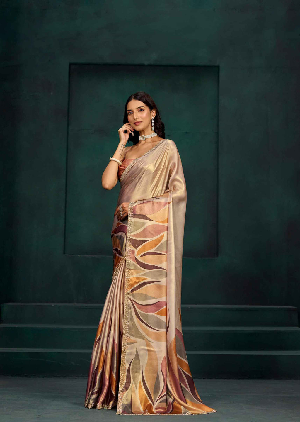 A woman models a draped saree with a metallic champagne-gold base and an abstract leaf-like pattern in bronze, brown, and orange tones, set against a dark green textured studio backdrop.