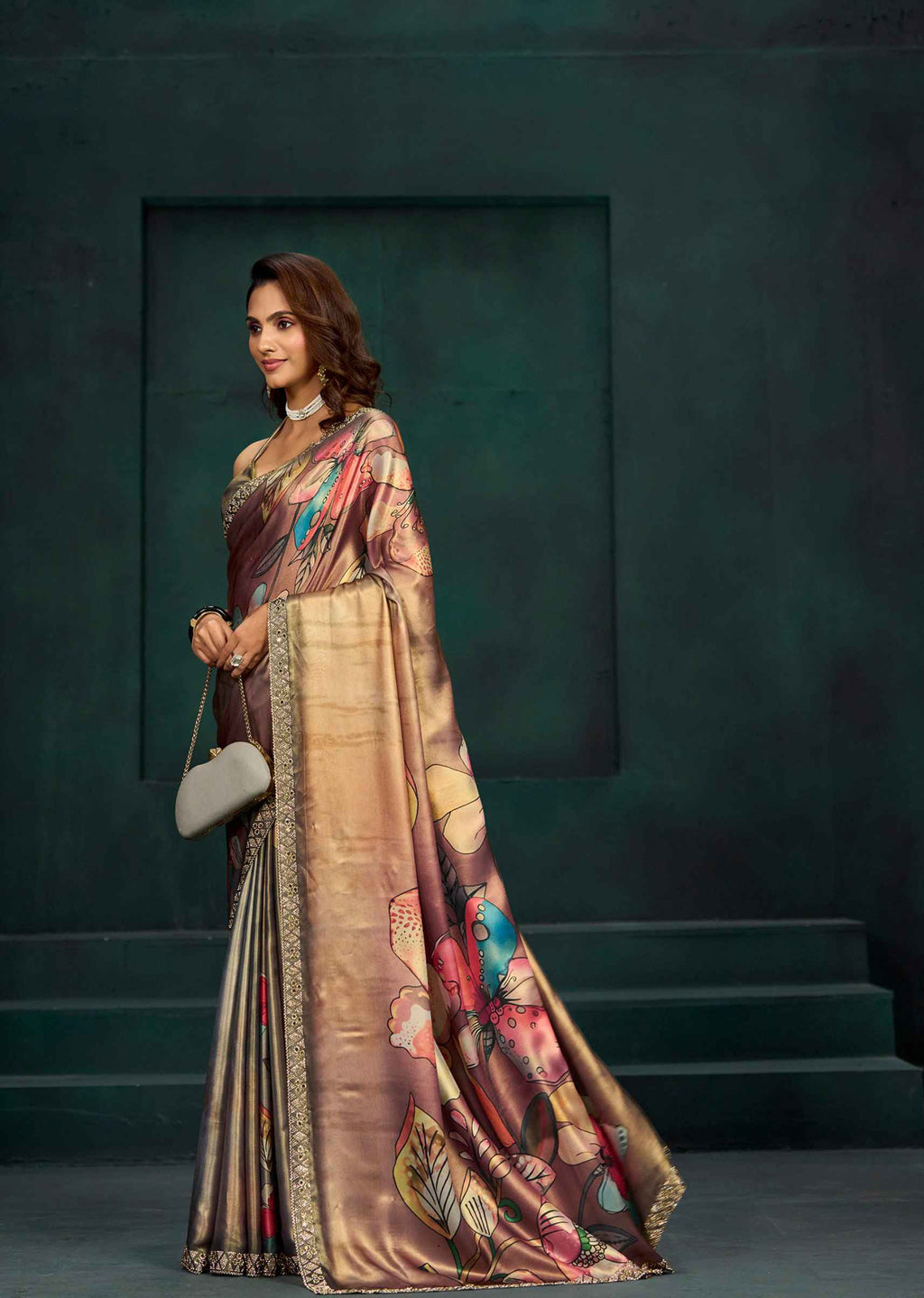 A woman models a luxurious bronze and gold saree with colorful floral patterns and a gold border, standing against a dark green textured studio backdrop.