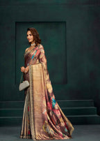 A woman models a luxurious bronze and gold saree with colorful floral patterns and a gold border, standing against a dark green textured studio backdrop.