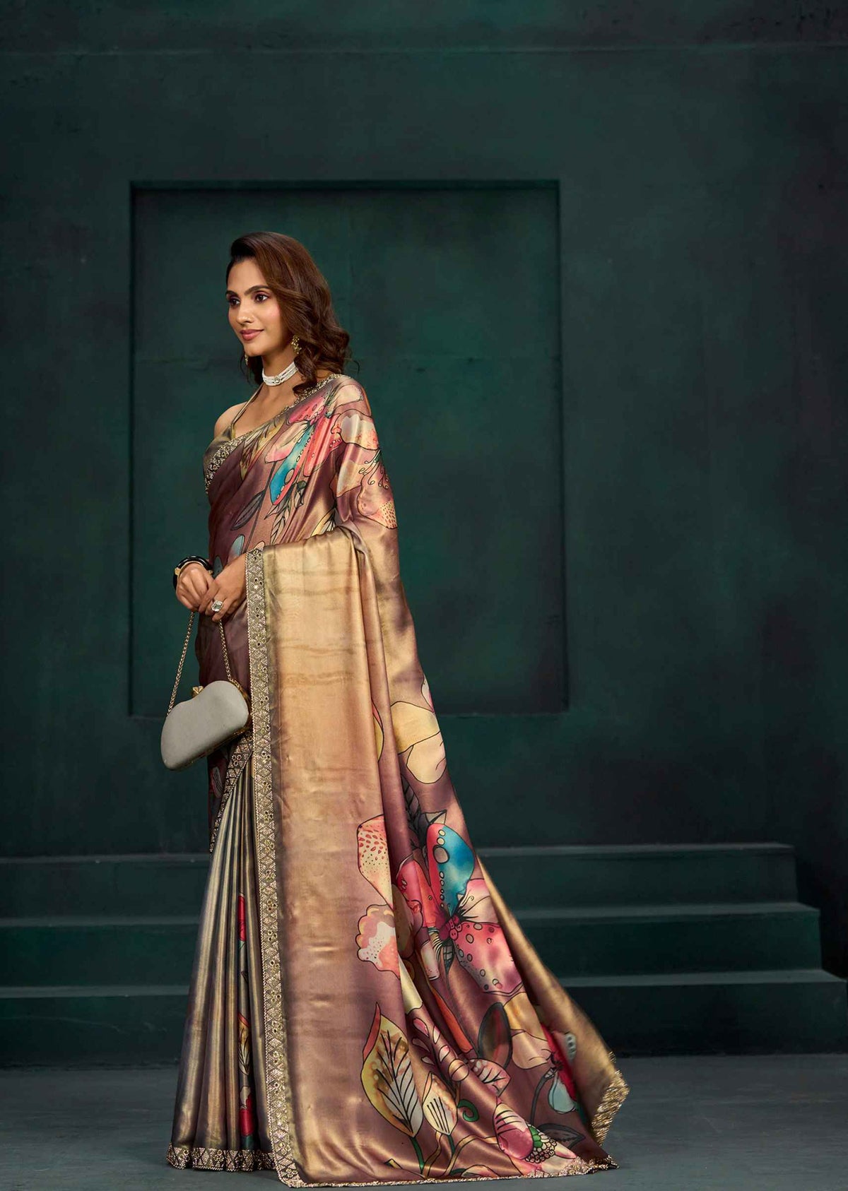 A woman models a luxurious bronze and gold saree with colorful floral patterns and a gold border, standing against a dark green textured studio backdrop.
