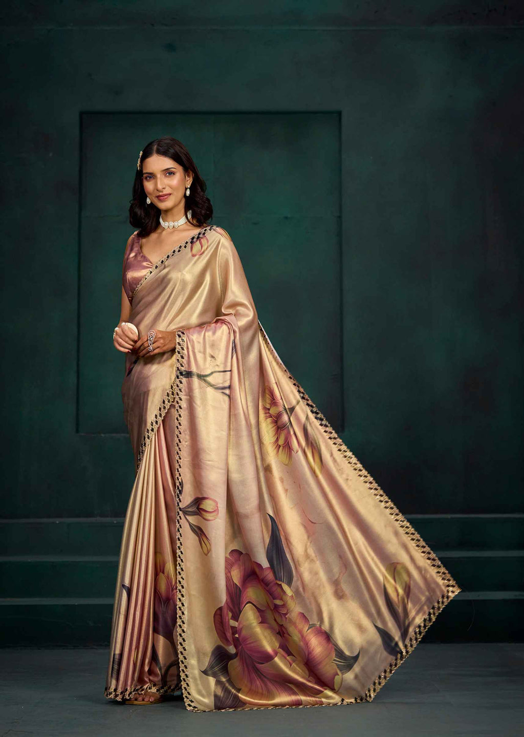 A woman models a metallic champagne-gold silk saree with a large floral print in pink, purple, and green, paired with a matching blouse, set against a dark green textured studio background.