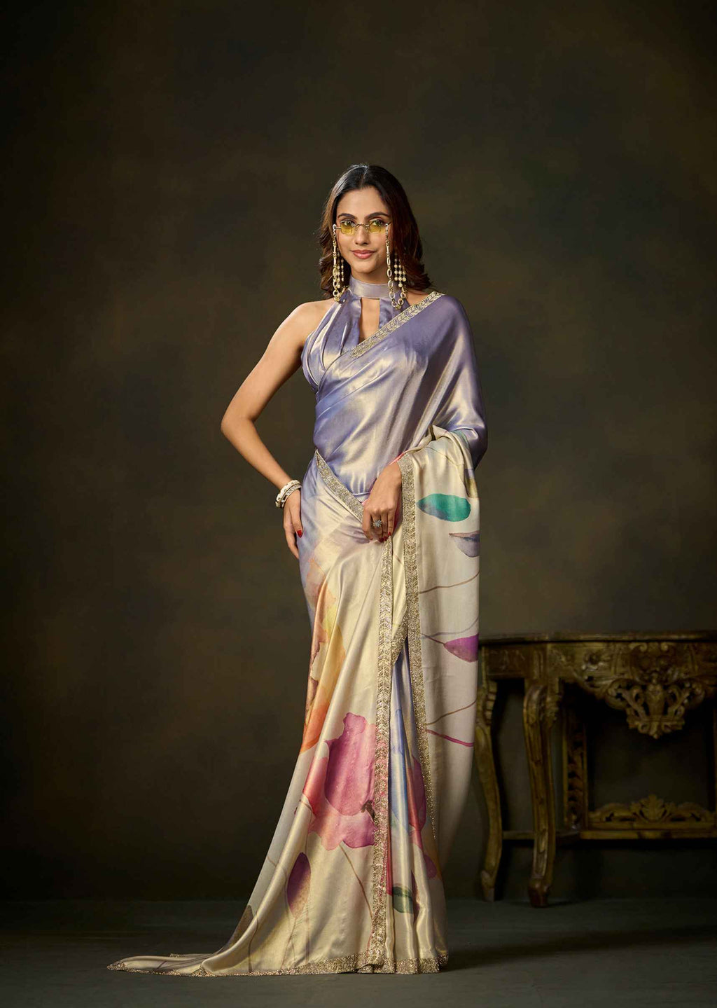 A woman models a draped satin saree in a studio setting. The saree has a lavender-purple upper drape and a cream-beige lower body with abstract pink, orange, and teal painted motifs, finished with a gold embroidered border.