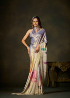 A woman models a draped satin saree in a studio setting. The saree has a lavender-purple upper drape and a cream-beige lower body with abstract pink, orange, and teal painted motifs, finished with a gold embroidered border.