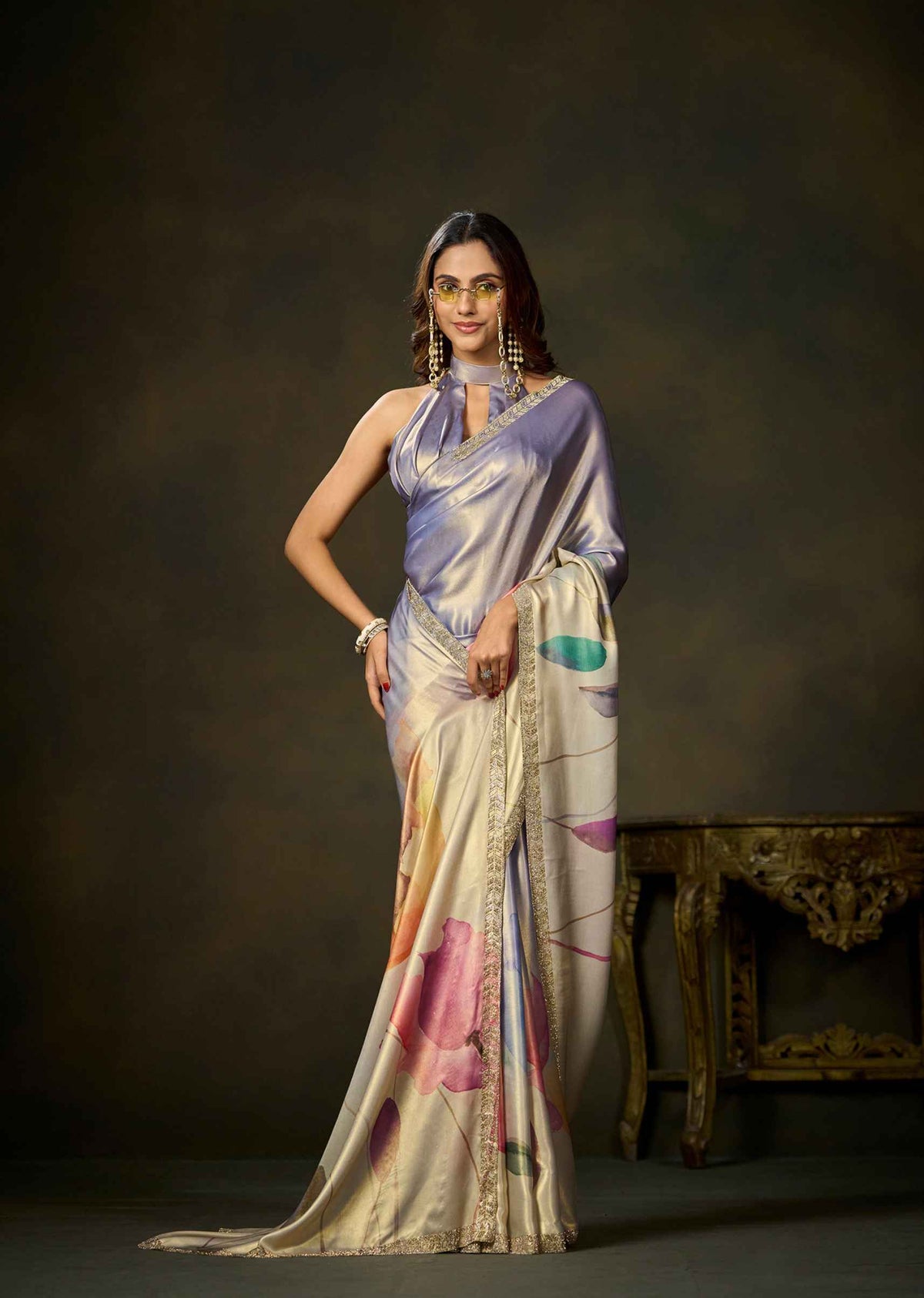 A woman models a draped satin saree in a studio setting. The saree has a lavender-purple upper drape and a cream-beige lower body with abstract pink, orange, and teal painted motifs, finished with a gold embroidered border.