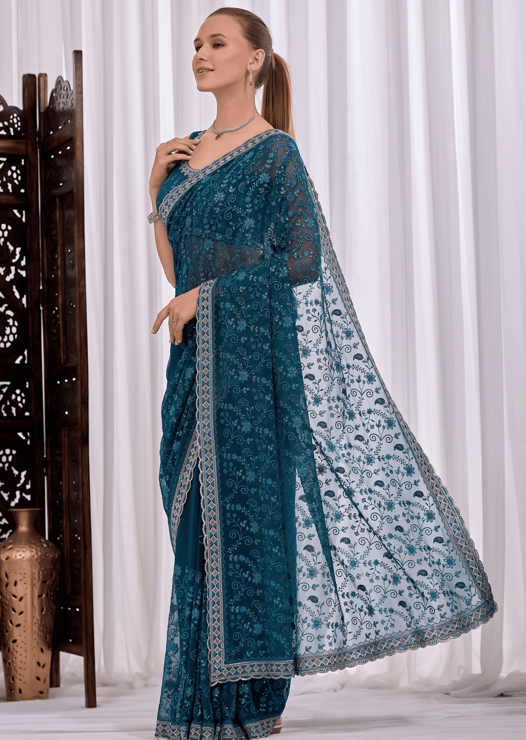 A woman models a teal blue embroidered saree with a matching dupatta, standing in front of white curtains with a decorative wooden screen on the left.