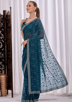 A woman models a teal blue embroidered saree with a matching dupatta, standing in front of white curtains with a decorative wooden screen on the left.