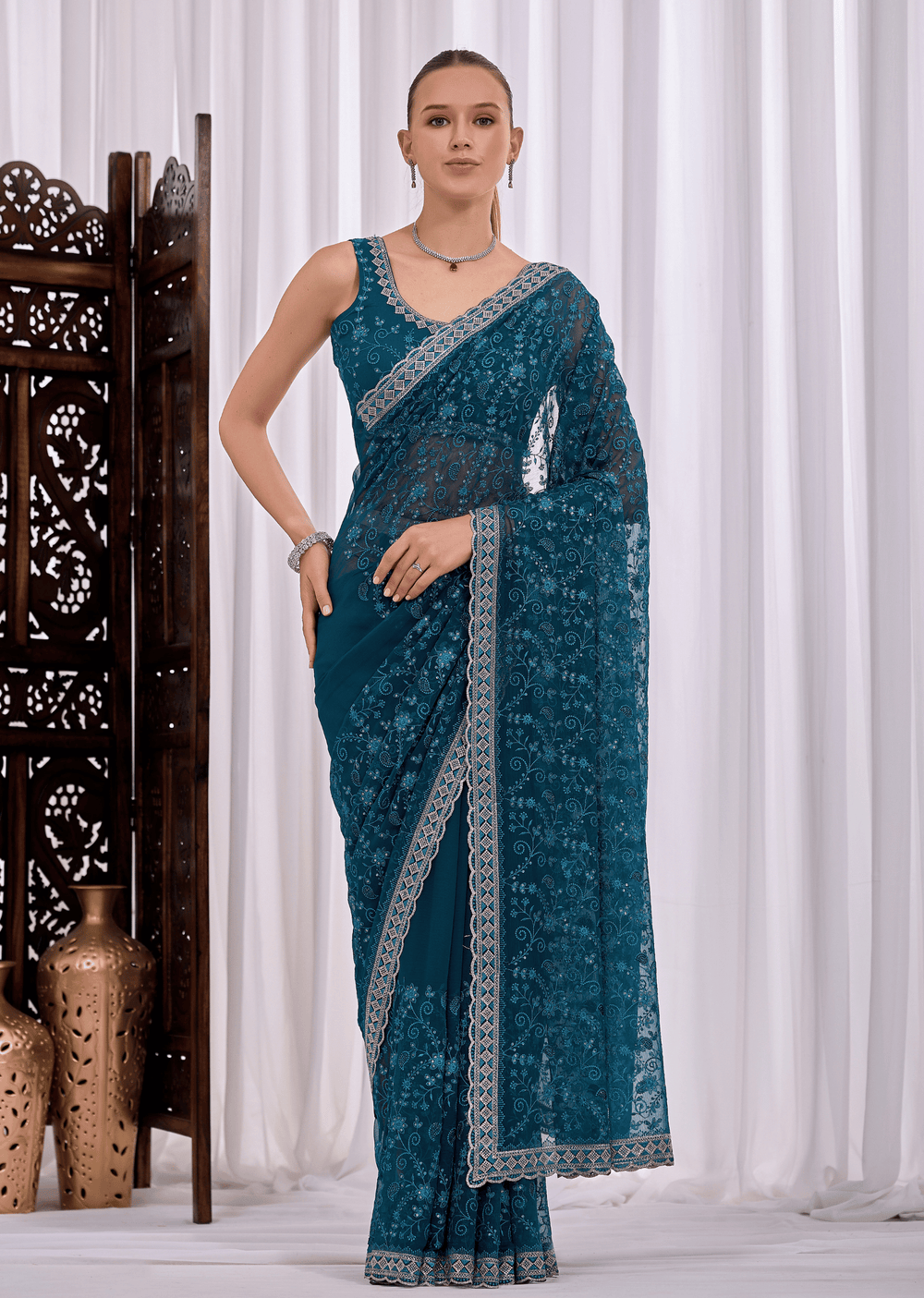 A woman modeling a teal blue saree with intricate silver floral embroidery and a matching embroidered border, standing in front of white curtains with a decorative wooden screen on the left.