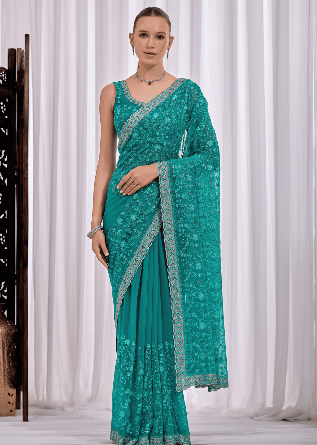 A woman modeling a teal green saree with intricate floral lace embroidery and a matching embroidered border, set against a white curtain backdrop.