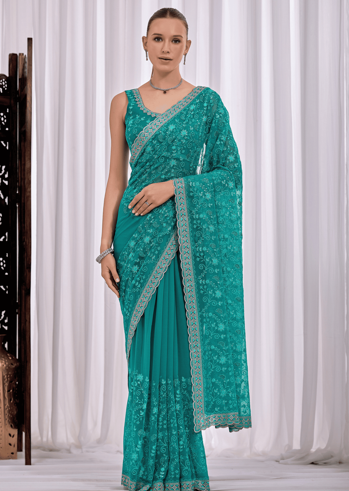 A woman modeling a teal green saree with intricate floral lace embroidery and a matching embroidered border, set against a white curtain backdrop.