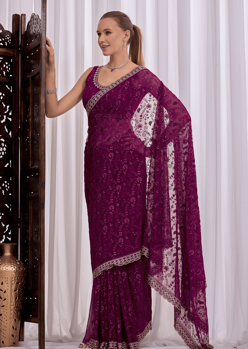 A woman models a deep purple georgette saree with intricate silver floral embroidery and a matching silver border, posed against a white curtain backdrop.