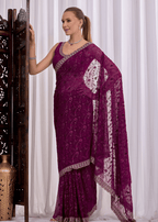 A woman models a deep purple georgette saree with intricate silver floral embroidery and a matching silver border, posed against a white curtain backdrop.