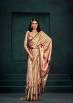 A woman models a shimmering champagne-gold saree with floral prints in pink, purple, and green, featuring a black and gold border. She is posed against a dark teal textured studio backdrop.