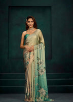 A woman models a traditional Indian saree with a sage green and beige body, featuring a light teal-green border with large white floral prints, set against a dark teal studio background.