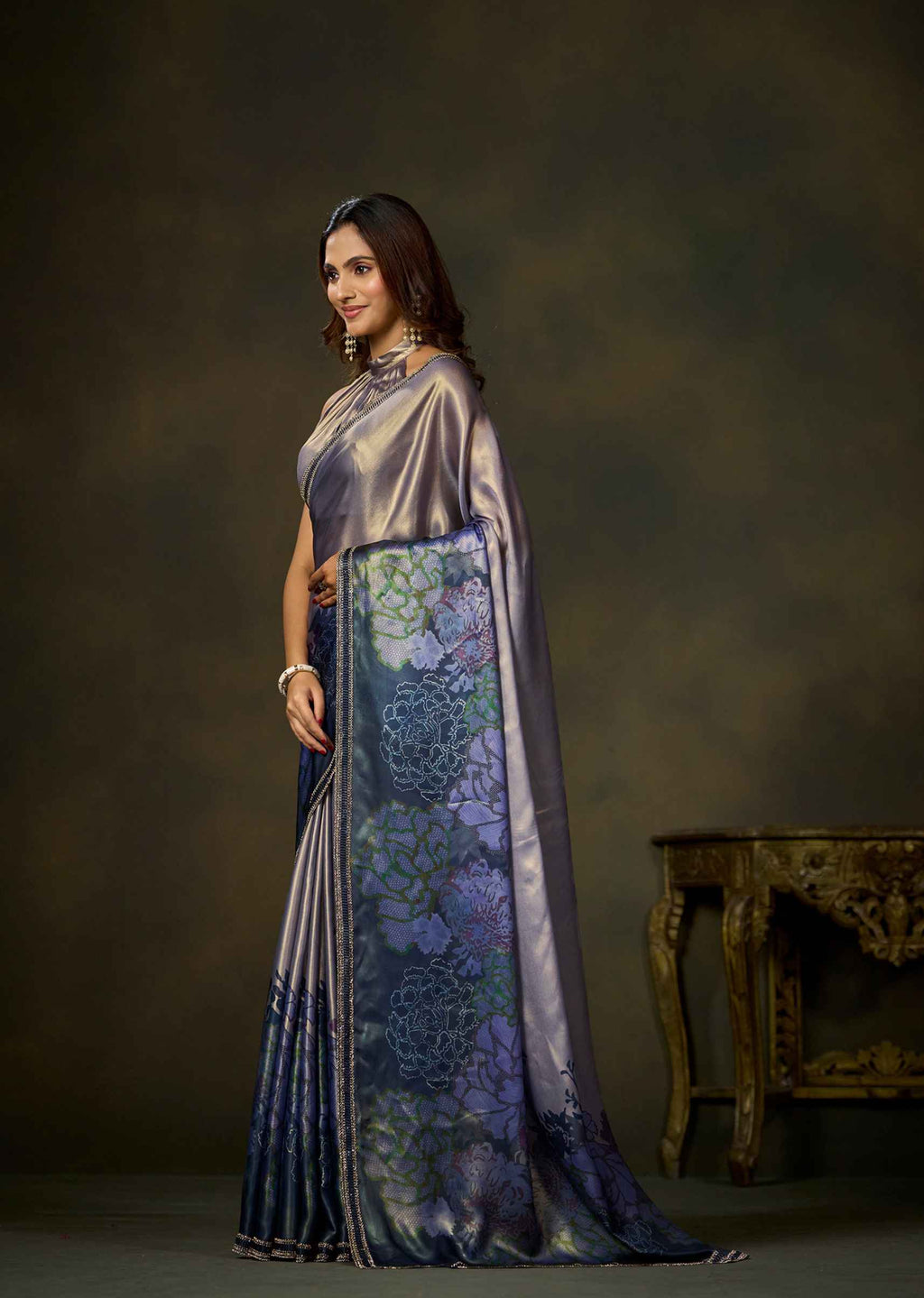 A woman modeling a traditional Indian saree in a studio setting. The saree features a lavender-purple satin body with a prominent border in deep blue and purple, decorated with floral patterns.