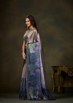 A woman modeling a traditional Indian saree in a studio setting. The saree features a lavender-purple satin body with a prominent border in deep blue and purple, decorated with floral patterns.