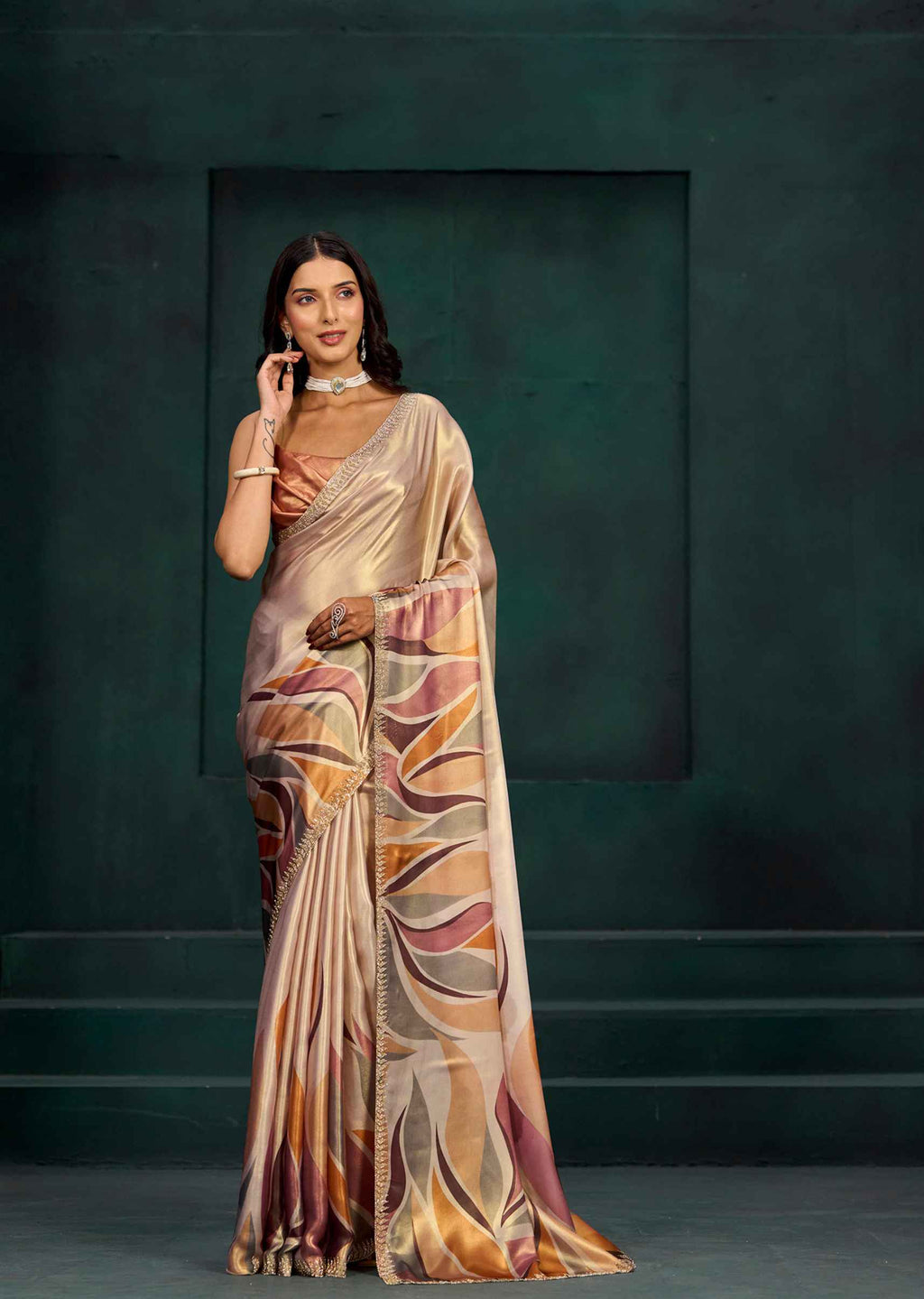 A woman models a draped satin saree with an abstract wave pattern in beige, gold, brown, and muted pink, set against a dark green textured studio backdrop.