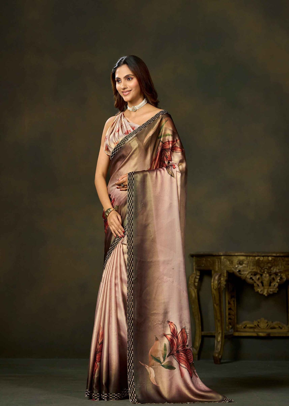 A woman models a draped satin saree in a studio setting. The saree is a shimmering champagne pink with a floral print in red and green on the pallu, and a black and gold patterned border.