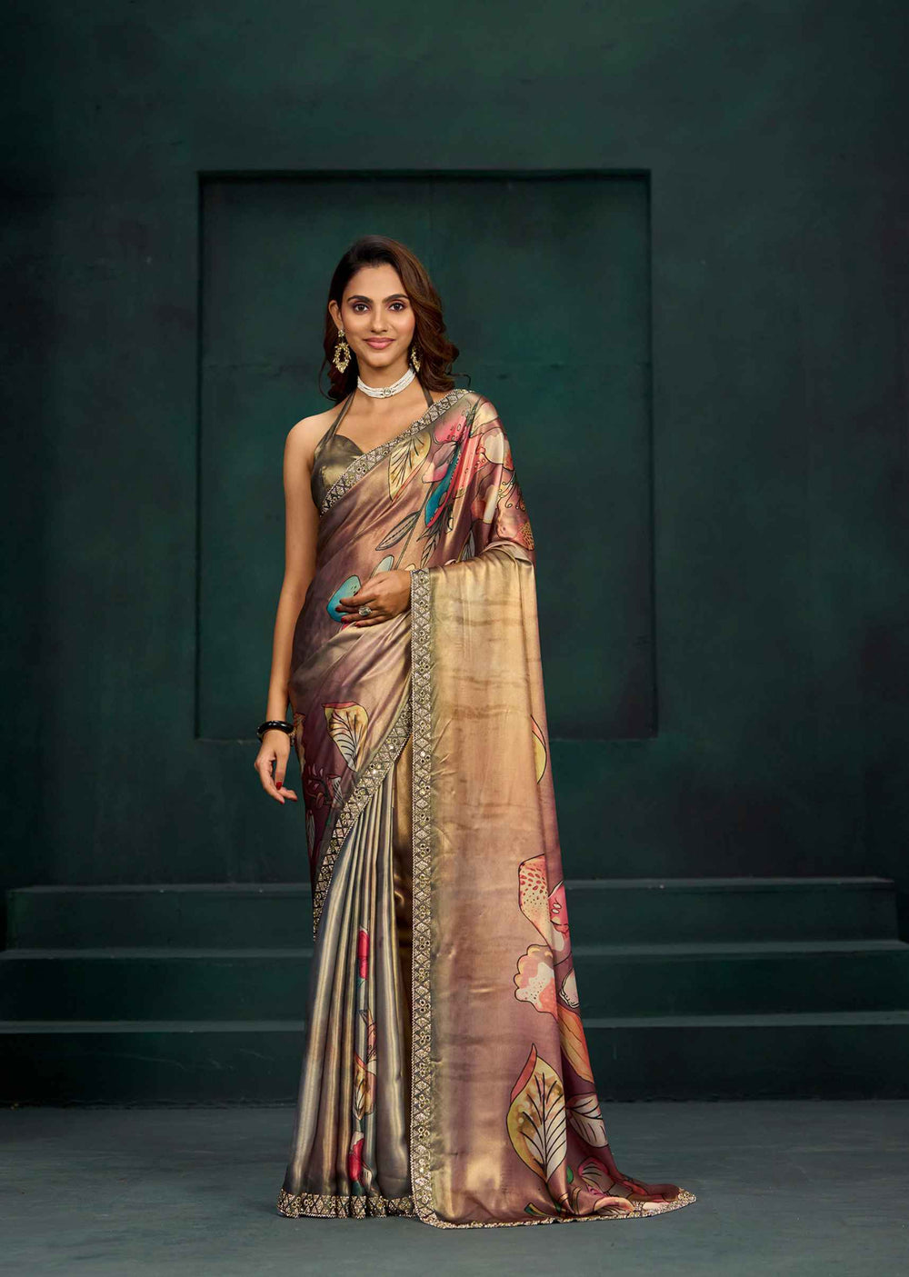 A woman modeling a draped saree in a studio setting with a dark green textured background. The saree has a metallic bronze and gold base with colorful floral and abstract patterns in pink, blue, and green, and is finished with a wide gold-trimmed border.
