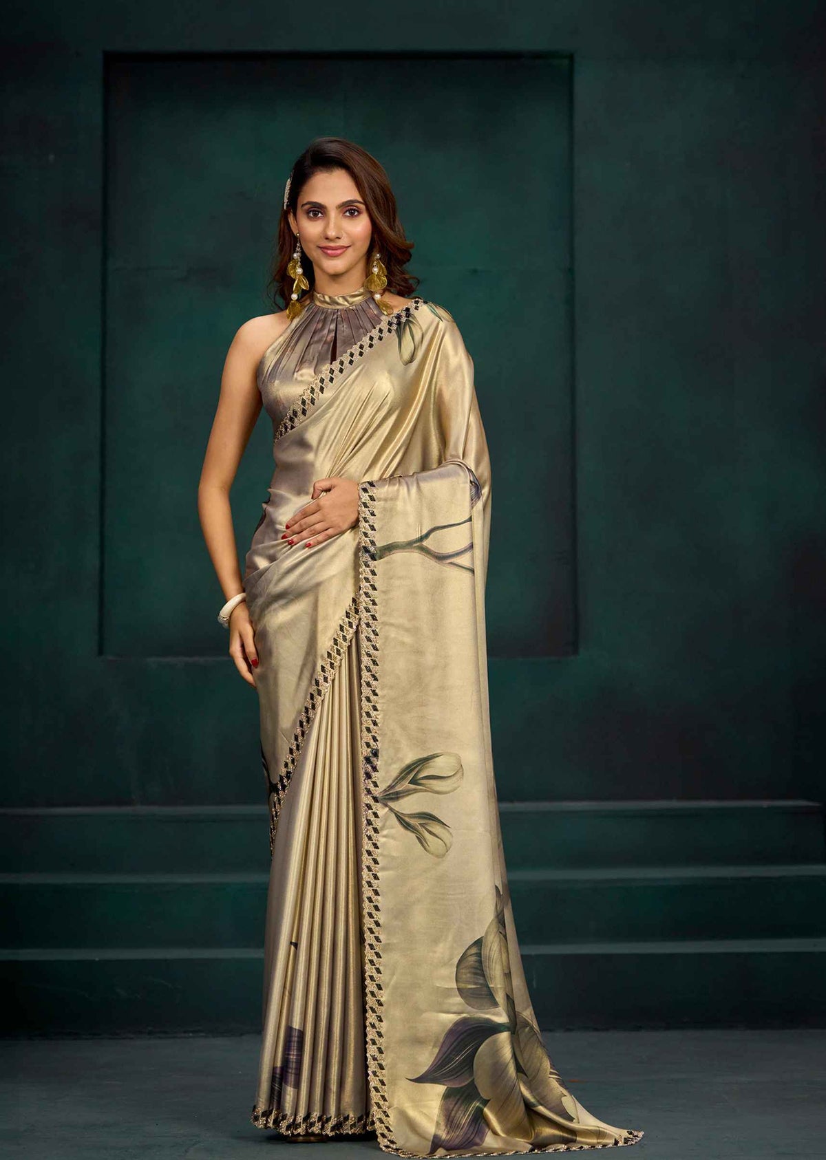 A woman models a metallic champagne-gold saree with black and green floral motifs and a black geometric border, set against a dark green studio backdrop.