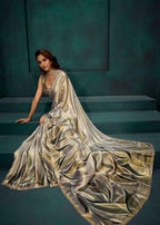 A woman wearing a flowing metallic champagne-gold sari with a matching sleeveless blouse, posed on dark teal steps against a matching teal wall.