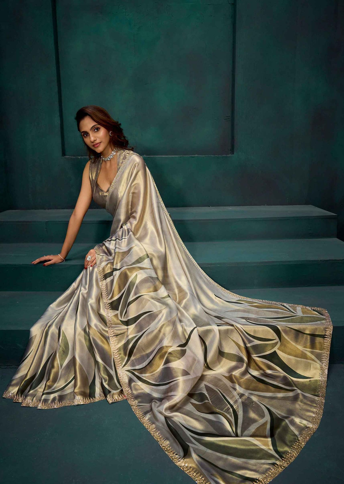 A woman wearing a flowing metallic champagne-gold sari with a matching sleeveless blouse, posed on dark teal steps against a matching teal wall.