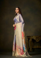A woman models a traditional Indian outfit featuring a lavender satin kurta paired with a beige dupatta decorated with a colorful watercolor floral print and gold zari borders, set against a dark studio background with an antique table.