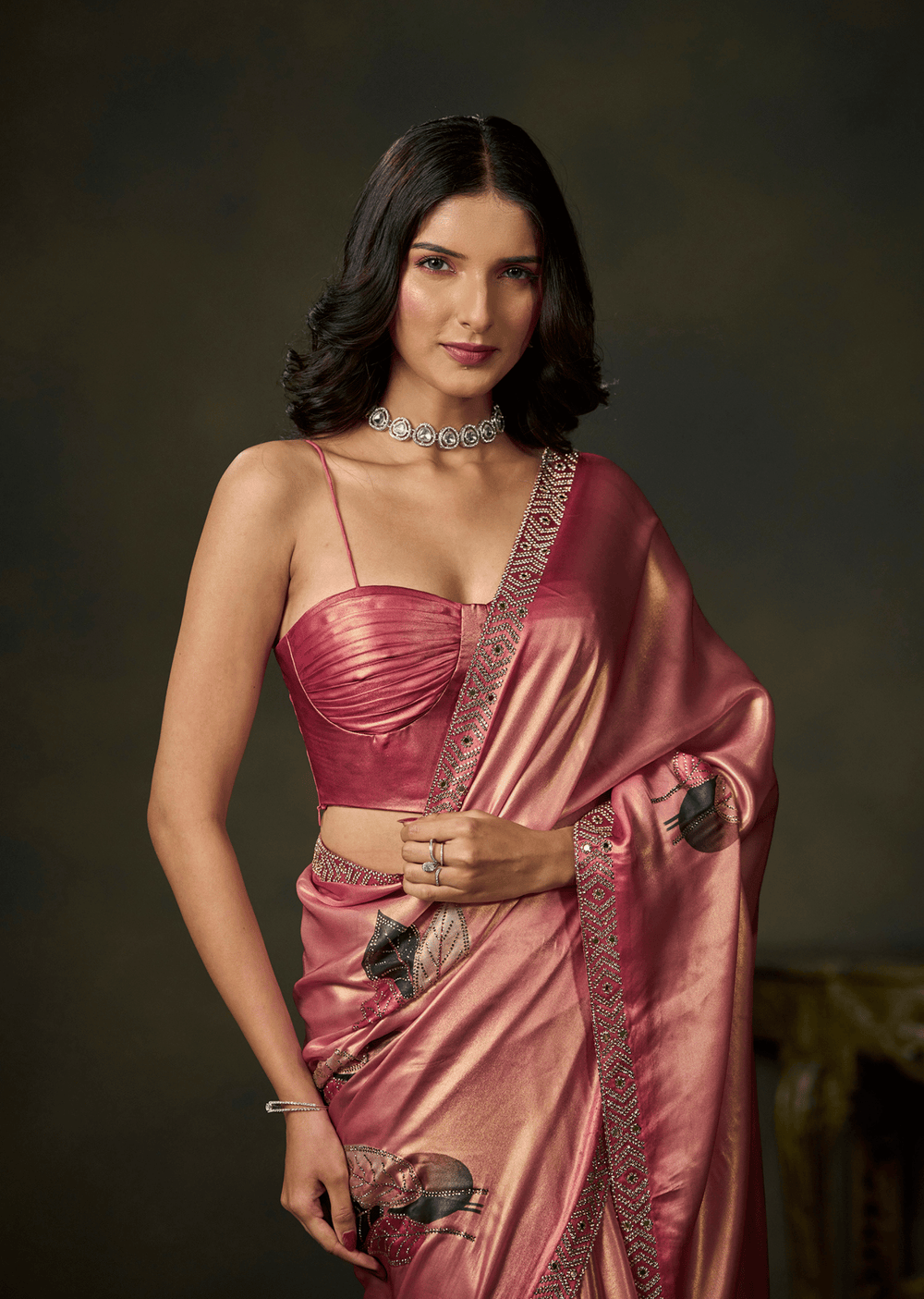 A woman modeling a metallic pink saree with a matching crop top and silver jewelry against a dark brown studio background.