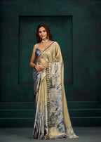 A woman models a traditional Indian saree with a metallic gold blouse and a draped saree body featuring a blue and gray watercolor floral pattern with a gold border. She stands against a dark green textured studio backdrop.