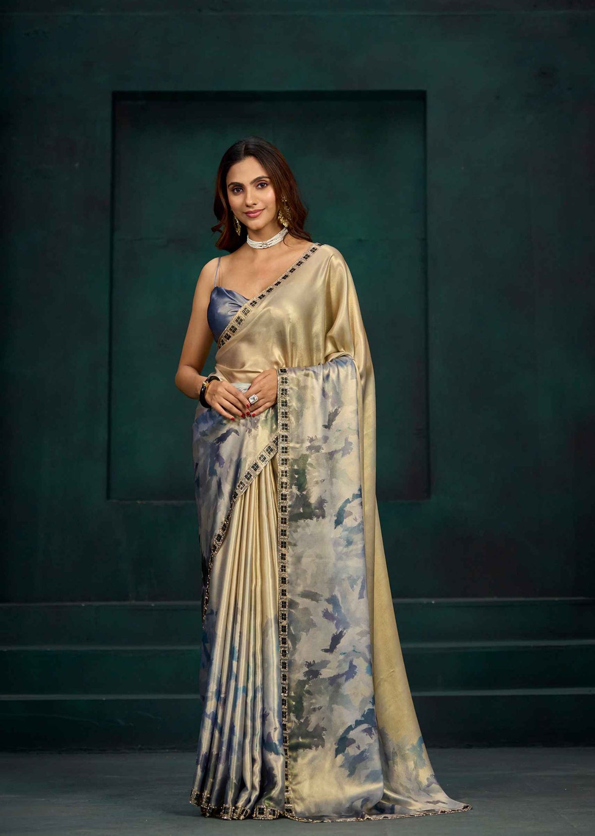 A woman models a traditional Indian saree with a metallic gold blouse and a draped saree body featuring a blue and gray watercolor floral pattern with a gold border. She stands against a dark green textured studio backdrop.