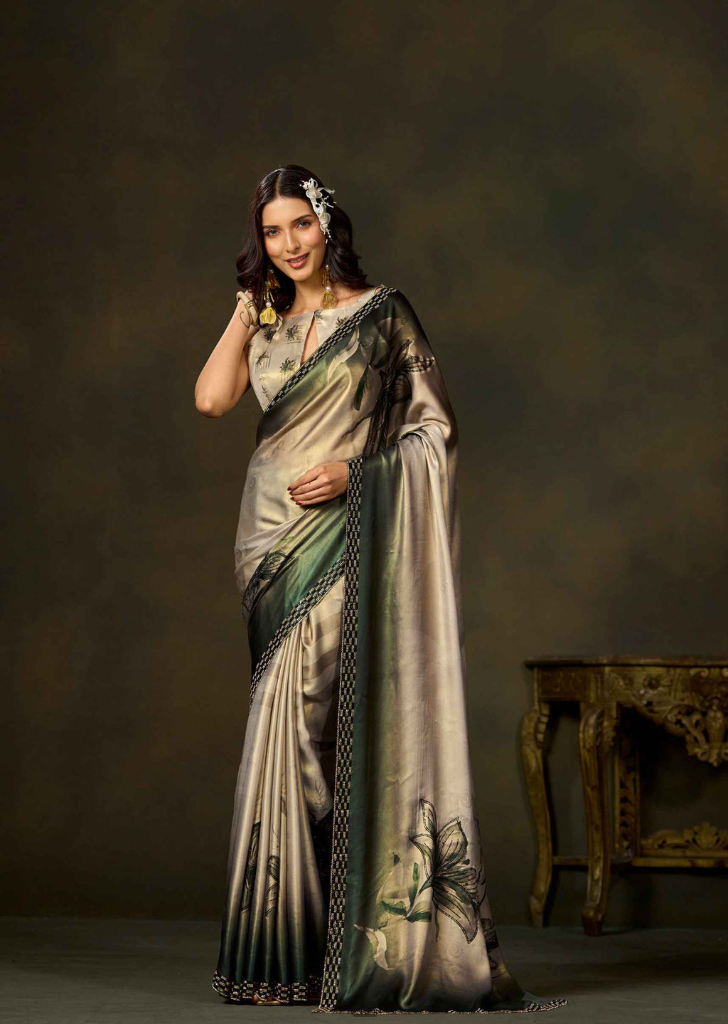 A woman modeling a metallic champagne-gold and olive green saree with black border detailing and floral motifs, posed against a dark brown studio backdrop with an ornate table in the background.