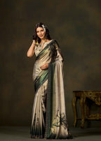 A woman modeling a metallic champagne-gold and olive green saree with black border detailing and floral motifs, posed against a dark brown studio backdrop with an ornate table in the background.