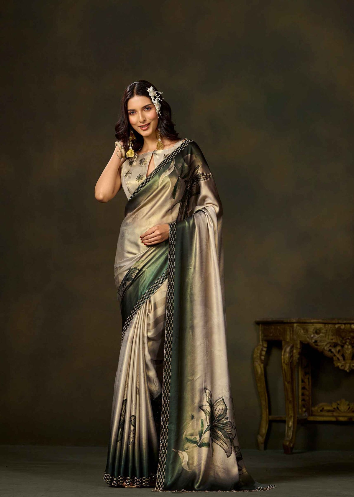 A woman modeling a metallic champagne-gold and olive green saree with black border detailing and floral motifs, posed against a dark brown studio backdrop with an ornate table in the background.
