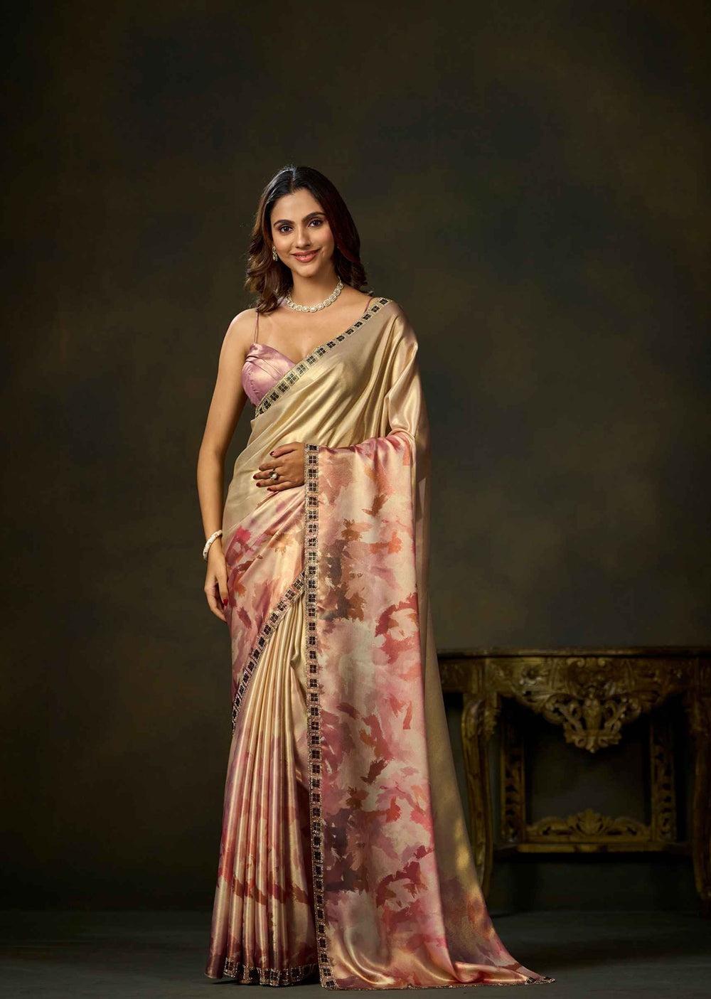 A woman models a traditional Indian saree with a metallic gold body, pink and rose watercolor floral print, and a black and gold border, set against a dark brown studio background.