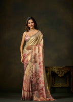 A woman models a traditional Indian saree with a metallic gold body, pink and rose watercolor floral print, and a black and gold border, set against a dark brown studio background.