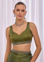 A woman modeling an olive green traditional-style two-piece outfit with a sleeveless crop top and matching skirt, both featuring a decorative silver patterned band at the waist and under the bust, against a white curtain background.