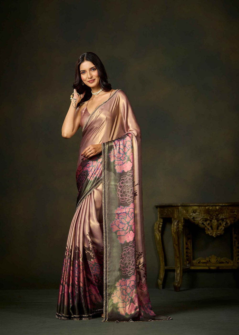 A woman models a draped satin saree with a metallic rose-gold sheen, featuring a pink and green floral pattern on the pallu and lower border, set against a dark studio background with an ornate table.