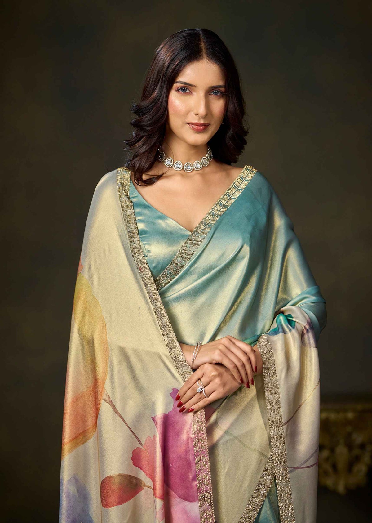 A woman modeling a light blue and beige printed silk saree with gold zari borders, styled with a silver choker necklace against a dark brown studio background.