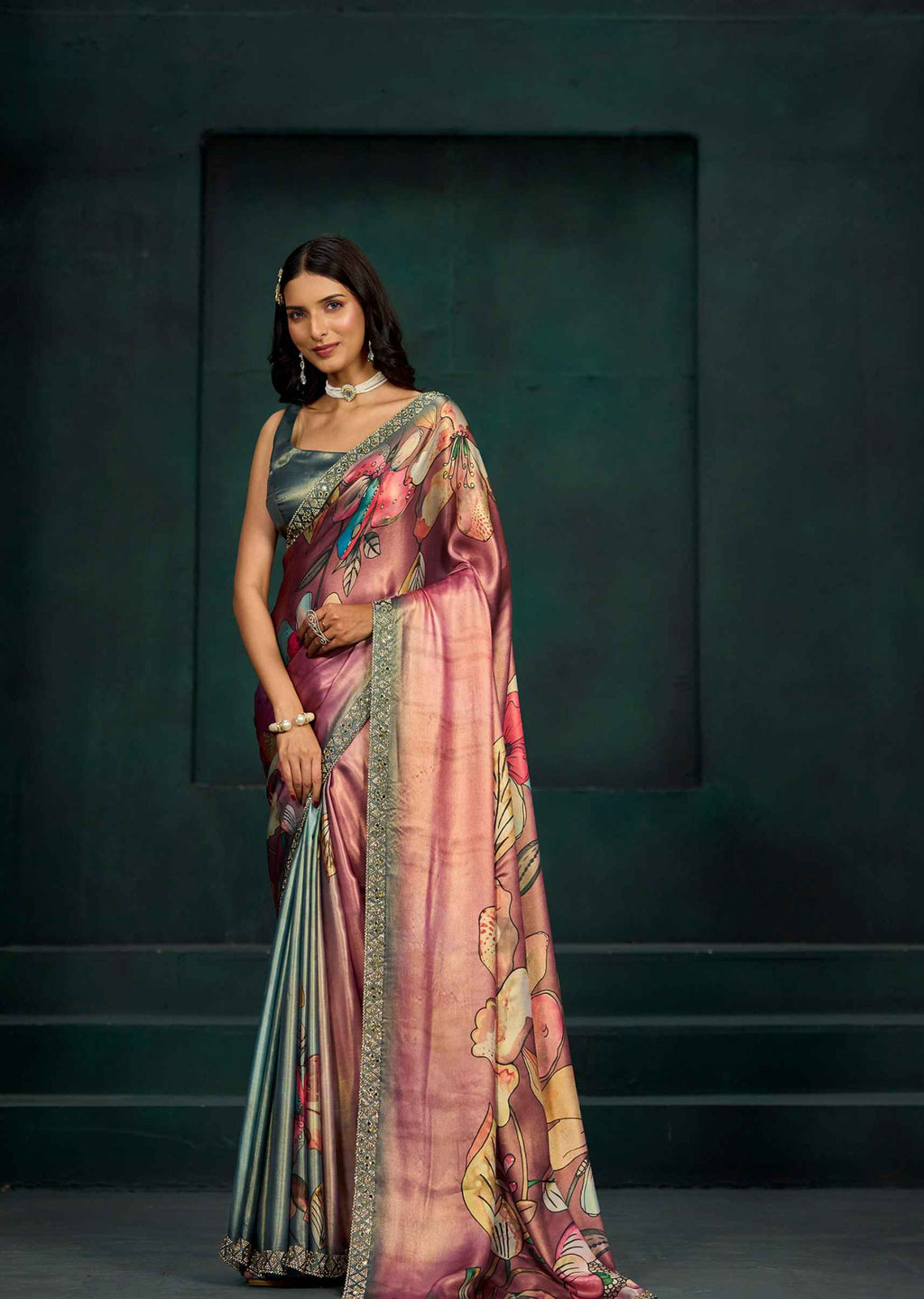 A woman models a pink and gold floral printed saree with a metallic blue-green blouse, standing against a dark textured studio backdrop.