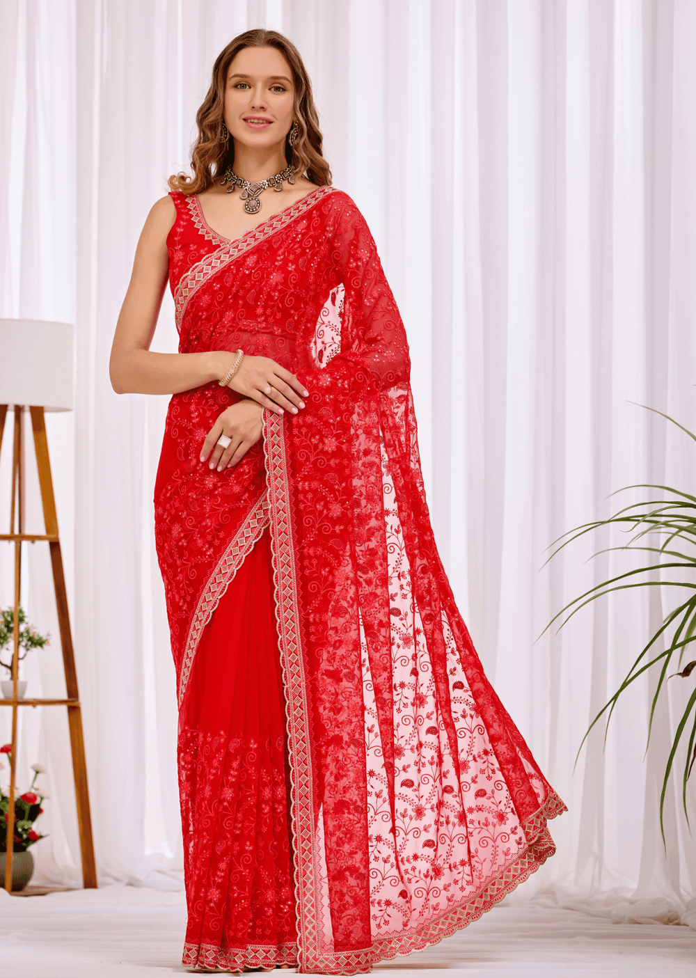A woman modeling a bright red saree with intricate floral lace embroidery and a matching red blouse, standing in front of white curtains with a plant and floor lamp visible in the background.