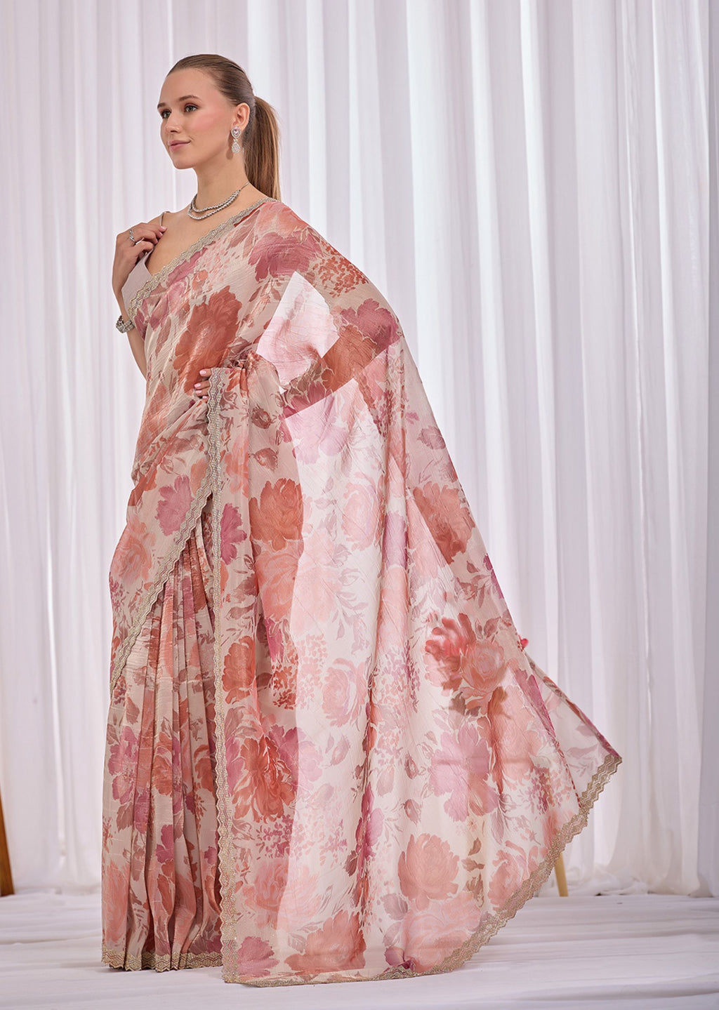 A woman modeling a light pink and coral floral printed saree with a matching dupatta, standing against a white curtain backdrop.