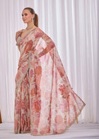 A woman modeling a light pink and coral floral printed saree with a matching dupatta, standing against a white curtain backdrop.