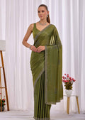 A woman modeling an olive green saree with silver embroidered borders and a matching sleeveless blouse, standing in front of a white curtain backdrop.