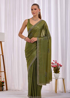 A woman modeling an olive green saree with silver embroidered borders and a matching sleeveless blouse, standing in front of a white curtain backdrop.