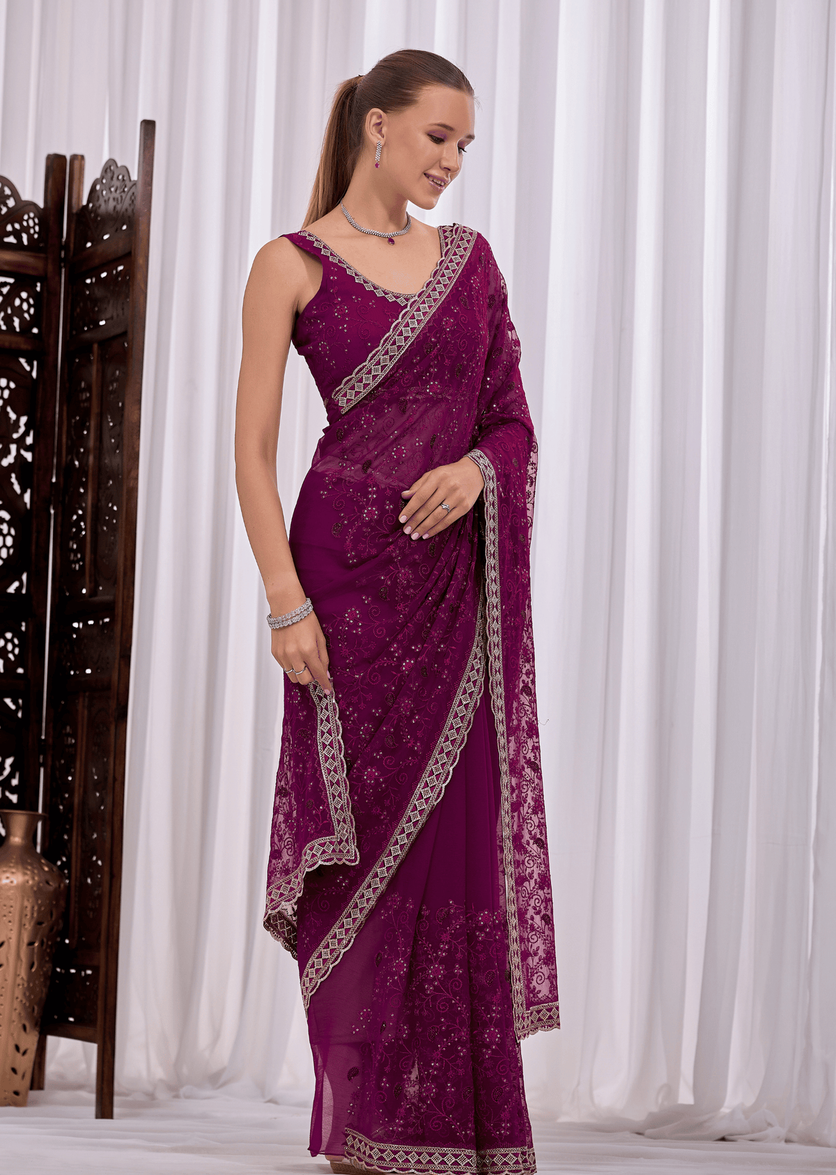 A woman modeling a deep purple embroidered saree with silver floral patterns and a matching border, standing in front of white curtains.